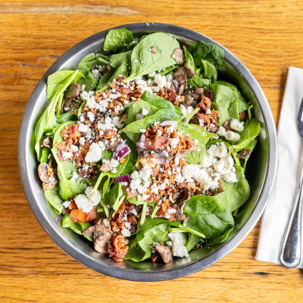 Spinach salad with bacon, feta cheese, mushrooms, and red onion in a metal bowl.
