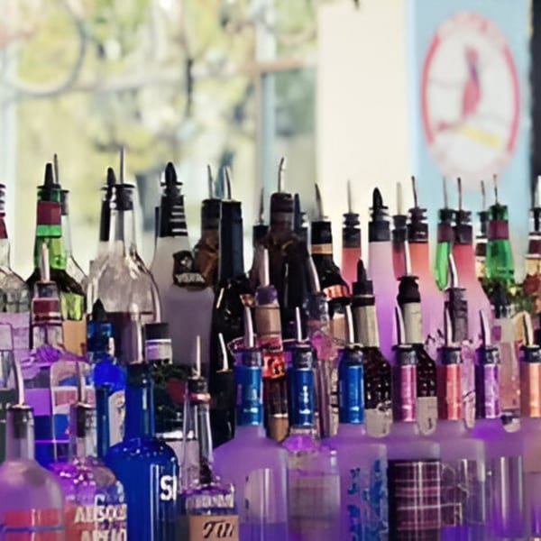 Numerous liquor bottles with pour spouts line a bar, illuminated by purple and pink light.